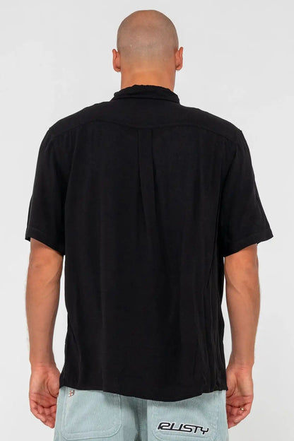 man wearing a black Rusty Mens Razor Blade Rayon Shirt and denim shorts on a pink background standing back on