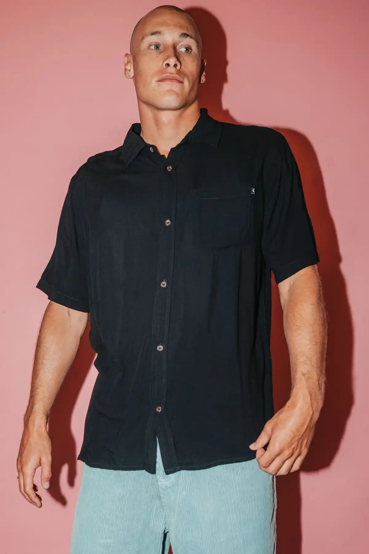 man wearing a black Rusty Mens Razor Blade Rayon Shirt and denim shorts on a pink background