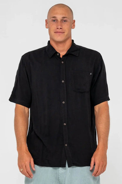 man wearing a black Rusty Mens Razor Blade Rayon Shirt and denim shorts on a white background