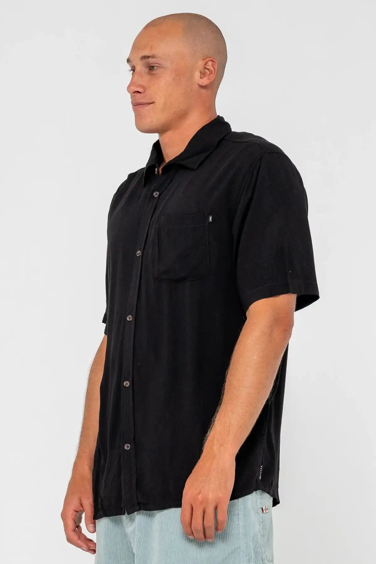 man standing side on wearing a black Rusty Mens Razor Blade Rayon Shirt and denim shorts on a white background