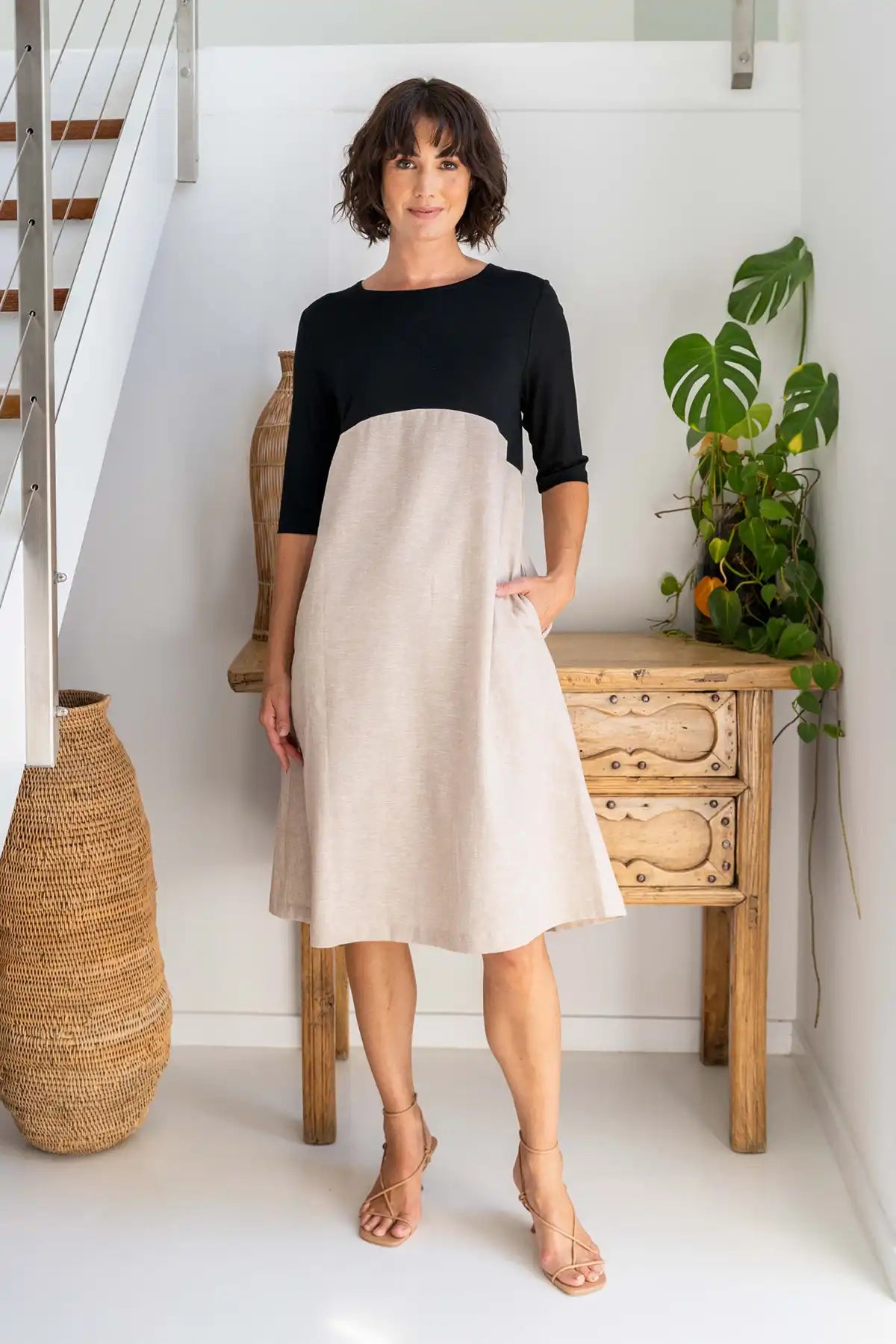 Woman wearing a See Saw Colour-Block Linen–Rayon Jersey Dress in Black/Natural standing in a home interior with a wooden table and plant.