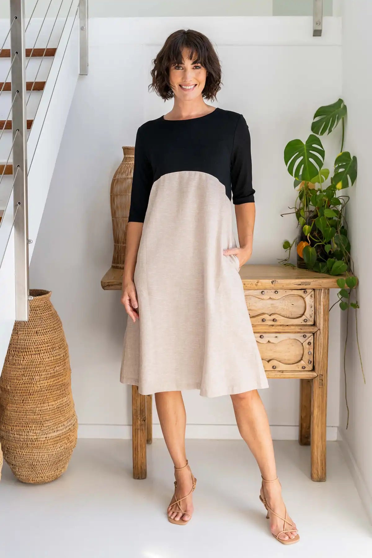 Woman wearing a See Saw Colour-Block Linen–Rayon Jersey Dress in Black/Natural standing in a home setting with a wooden side table and plant.