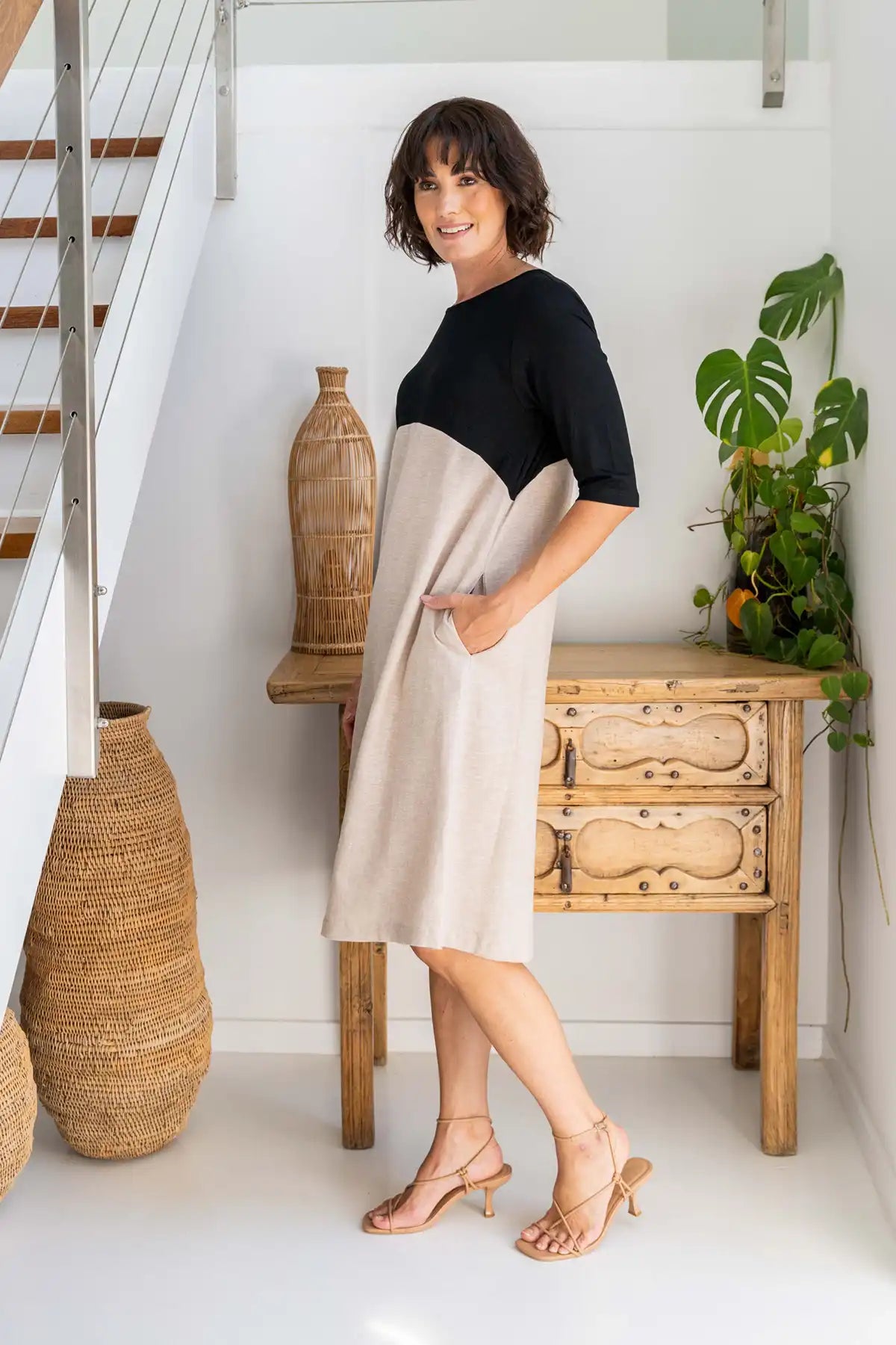 Woman wearing a See Saw Colour-Block Linen–Rayon Jersey Dress in Black/Natural standing side on in a room with wooden furniture and plants.