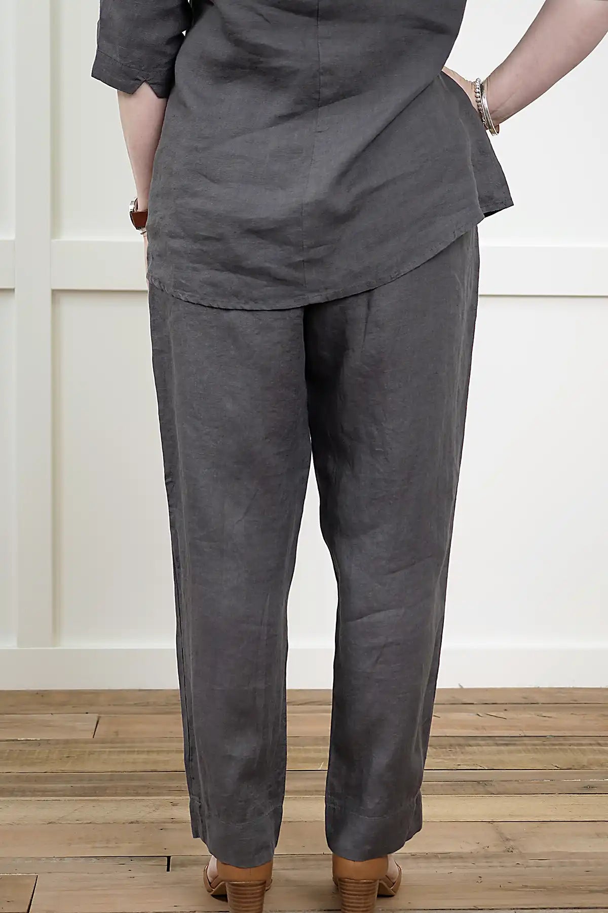 Person wearing a See Saw Linen 7/8 Flat Front Pant and cowl neck top in Charcoal  from behind and a top on a wooden floor.