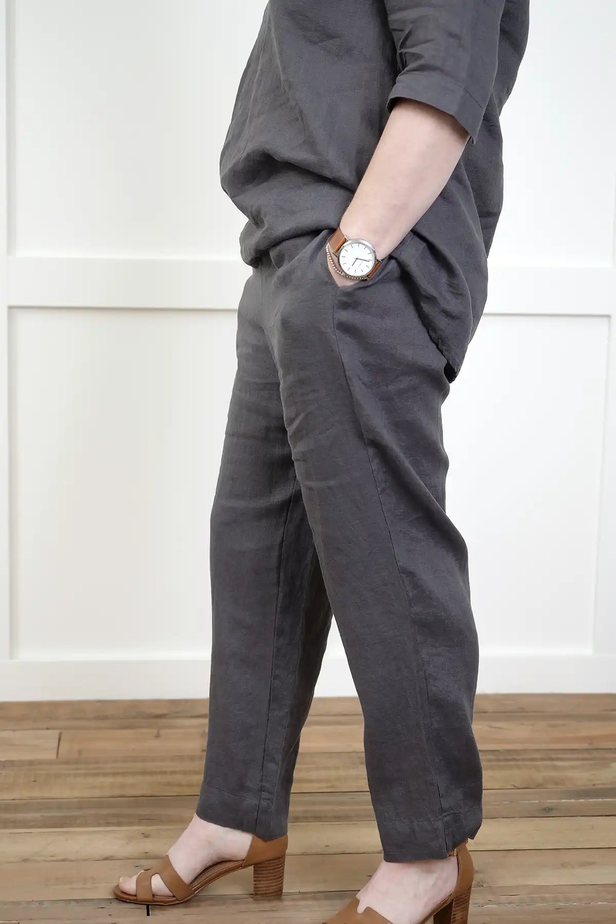 Person wearing See Saw Linen 7/8 Flat Front Pant and cowl neck top in Charcoal  and brown sandals side on against a white wall.