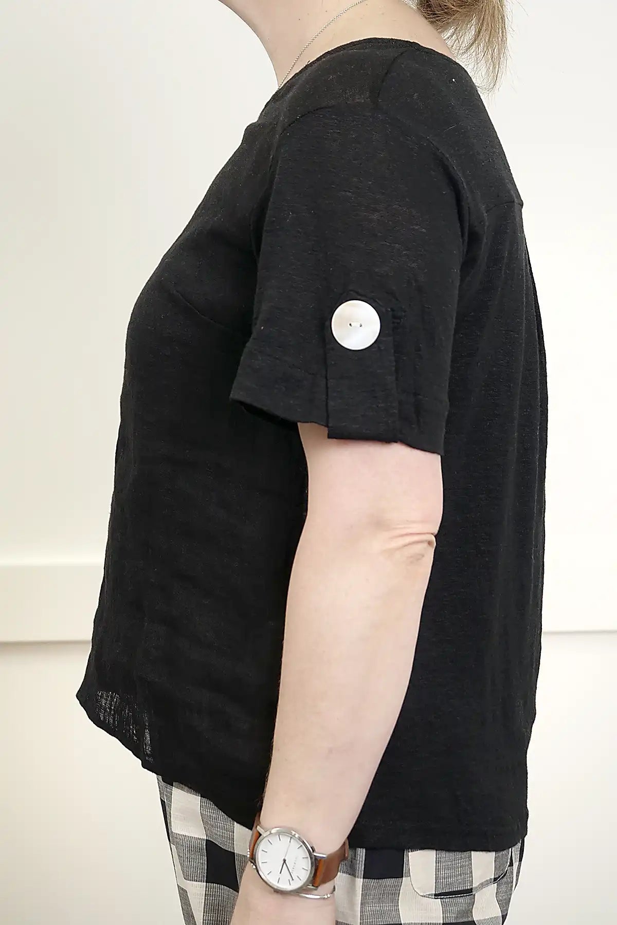 Person wearing aSee Saw Linen Mixed Texture Top in Black detailed view of the sleeve with a white button on a plain background