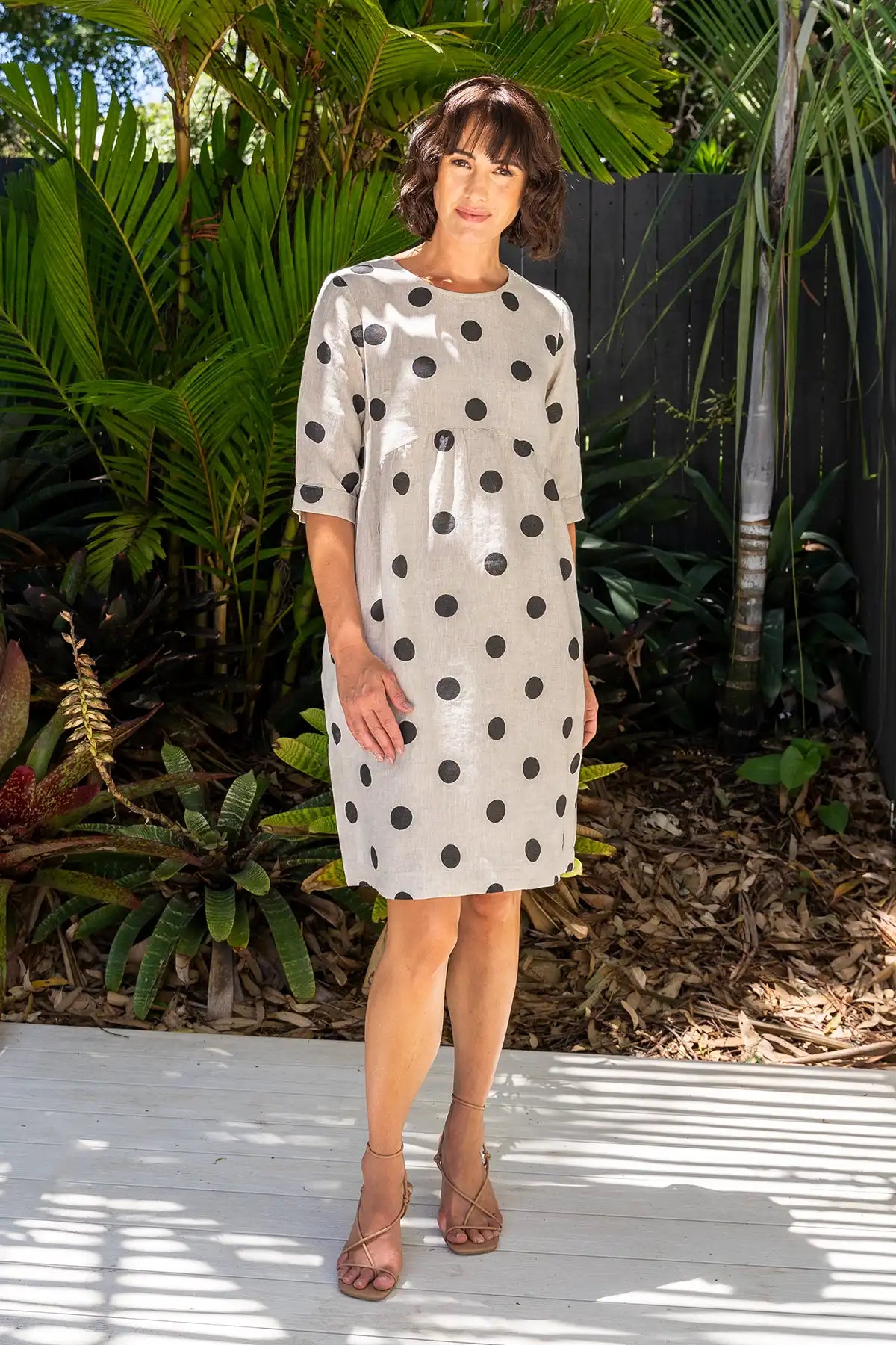 Woman wearing a See Saw Linen Spot Button-Back Dress in Black on Flax standing outdoors with plants in the background