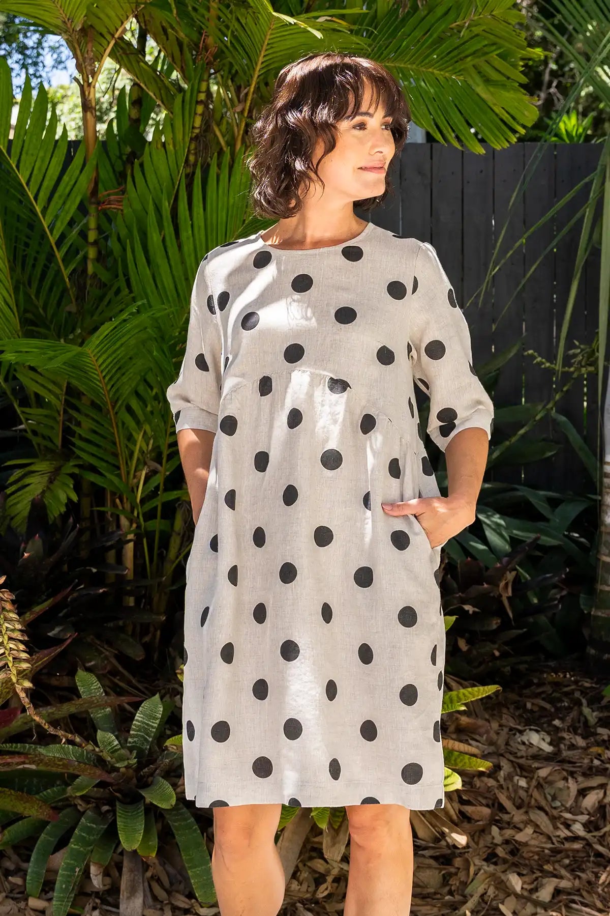 Woman wearing a See Saw Linen Spot Button-Back Dress in Black on Flax standing outdoors with greenery in the background
