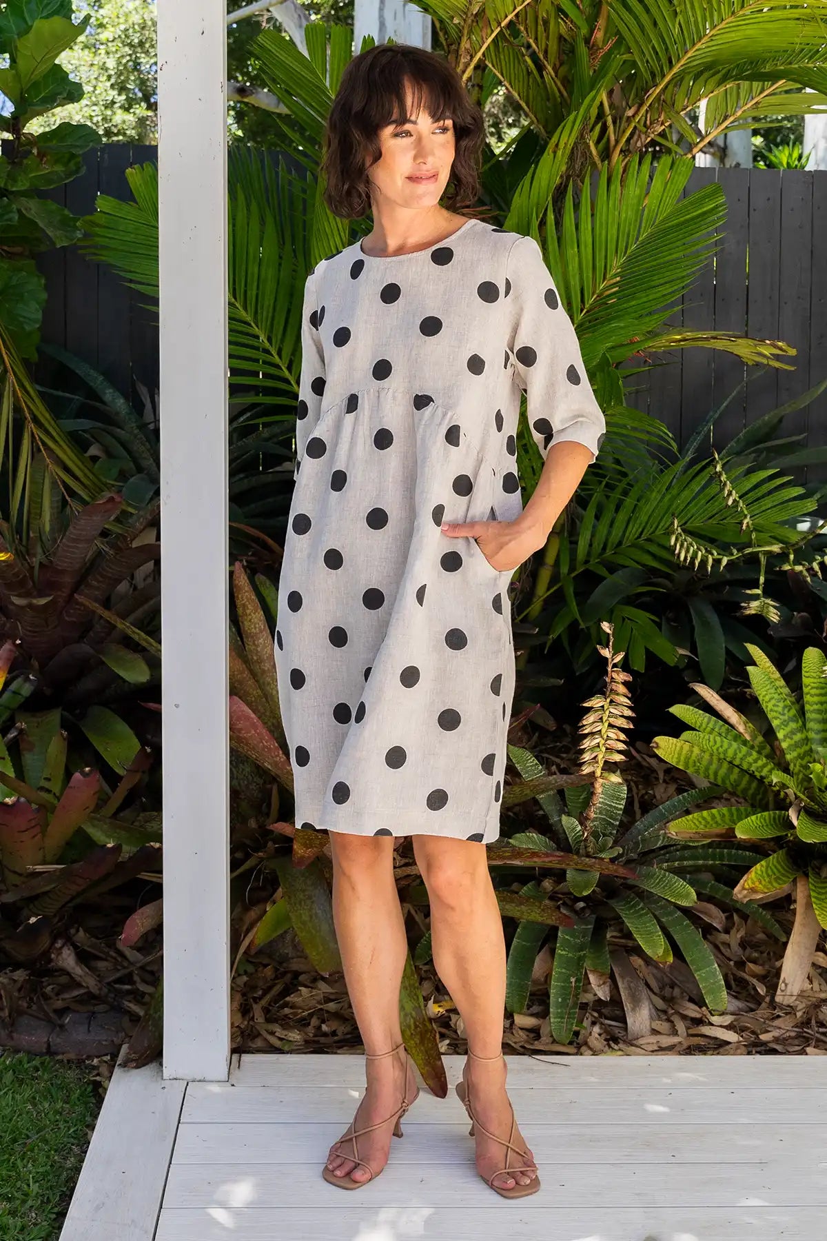 Woman wearing a See Saw Linen Spot Button-Back Dress in Black on Flax standing front on with green foliage in the background