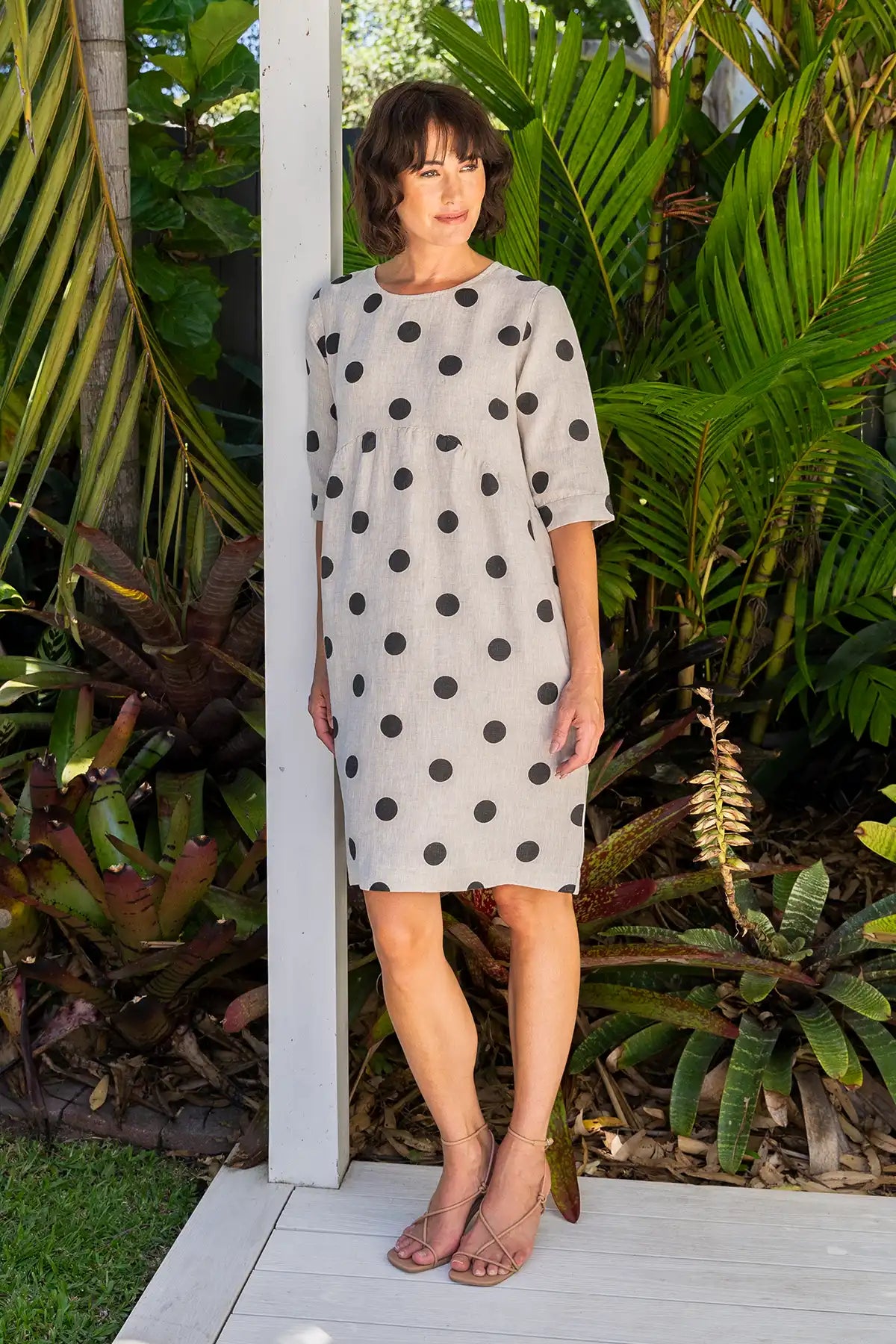 Woman wearing a See Saw Linen Spot Button-Back Dress in Black on Flax leaning on a post with greenery in the background