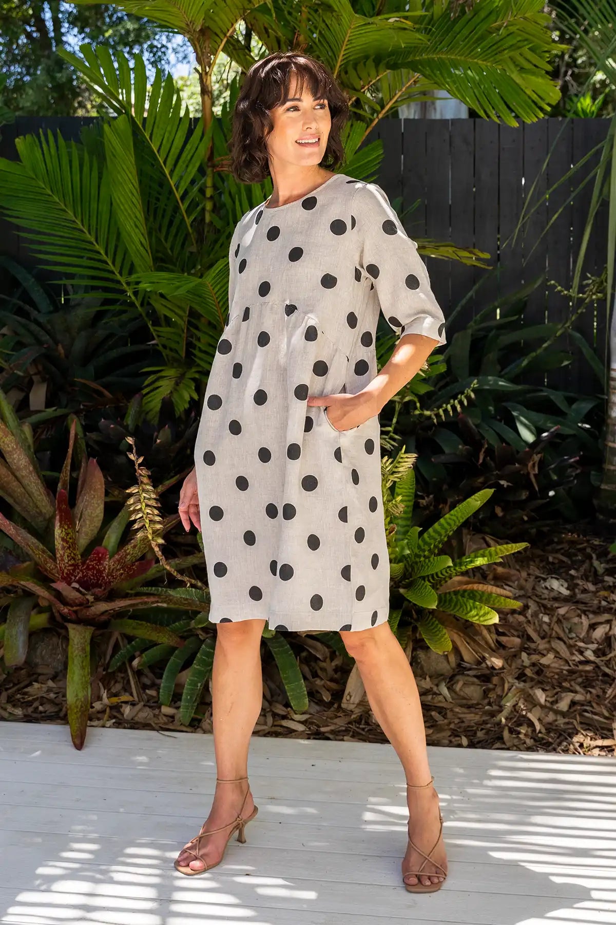 Woman wearing a See Saw Linen Spot Button-Back Dress in Black on Flax with hand in pocket standing outdoors with greenery in the background