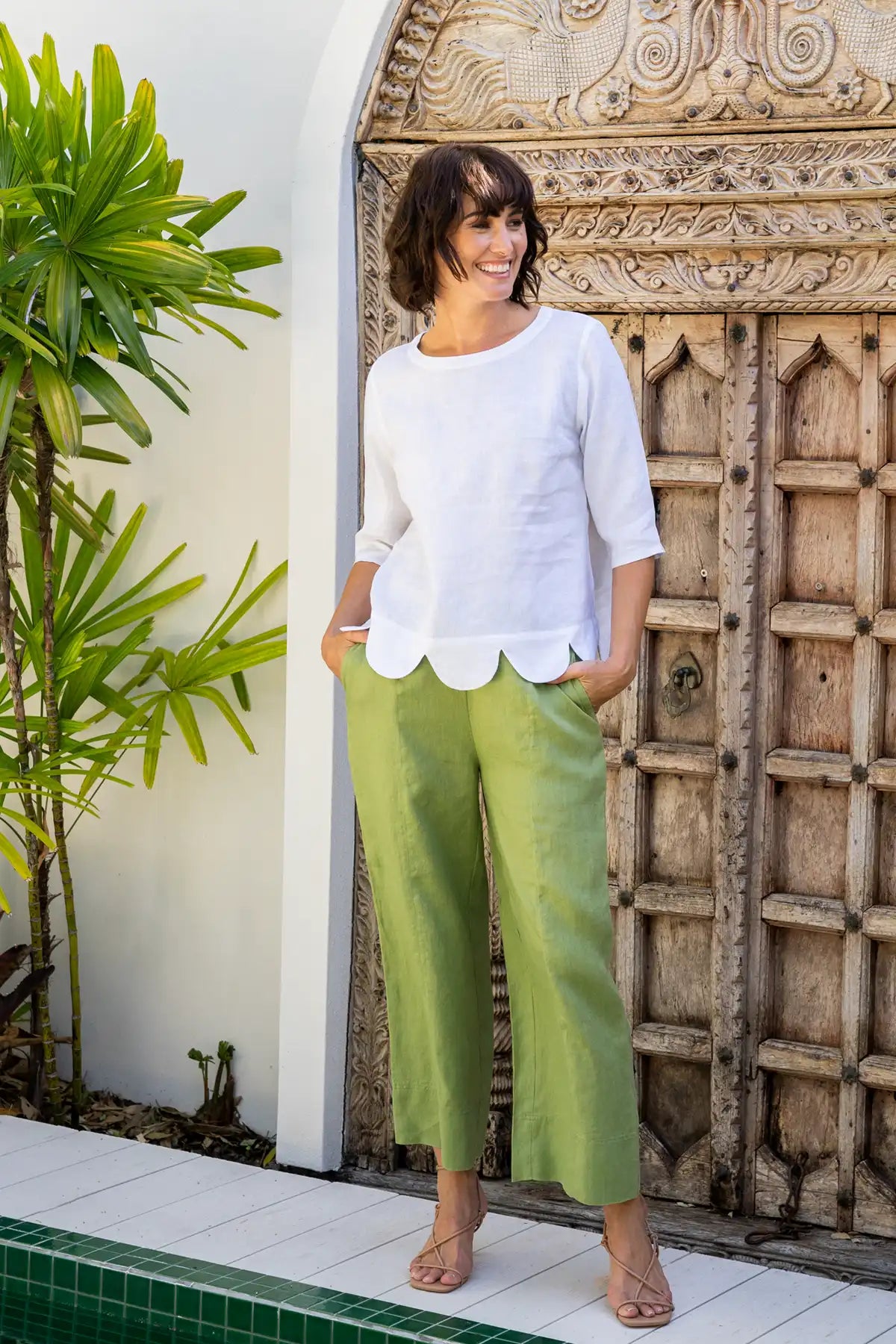 Woman in a white top and green pants standing in front of a decorative wooden door.
