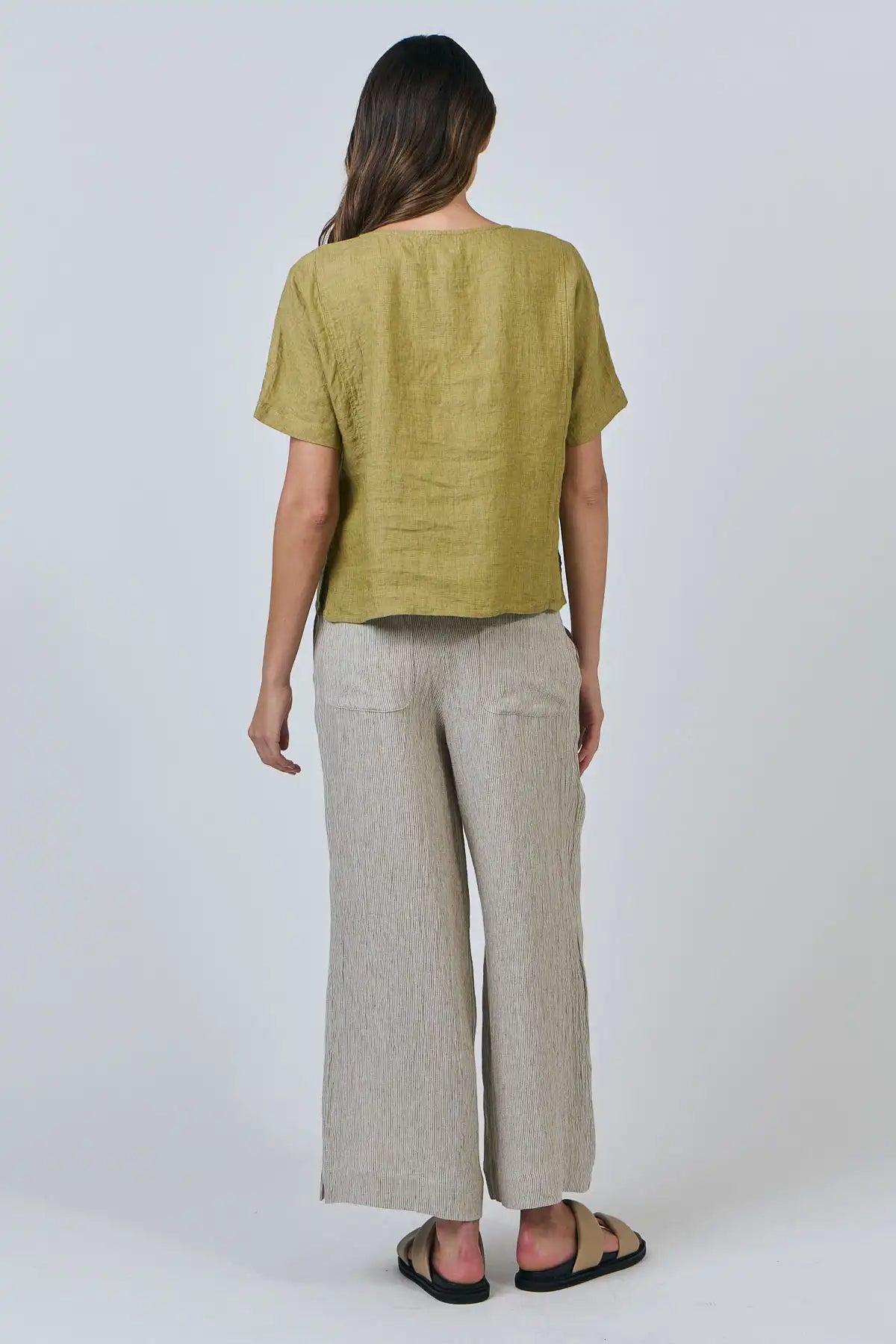 Woman wearing a Solace Linen Top in green Bamboo and beige pants on a white background full model back view