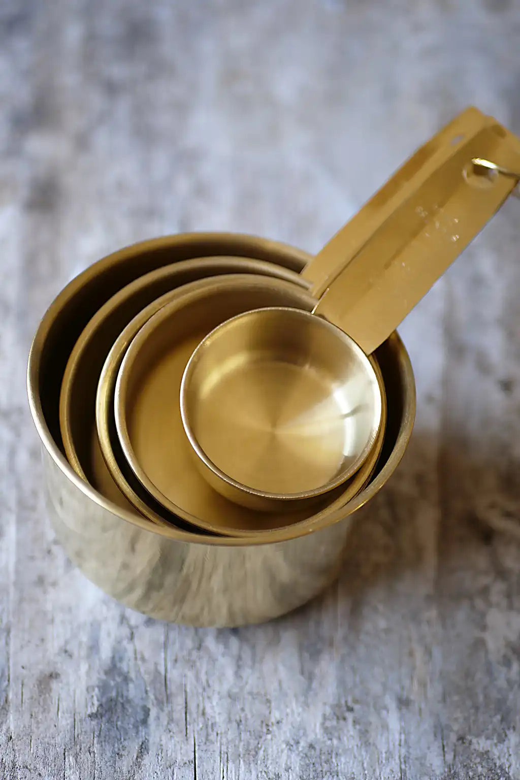 A set of nested brass measuring cups with long handles, arranged on a rustic gray wooden surface.
