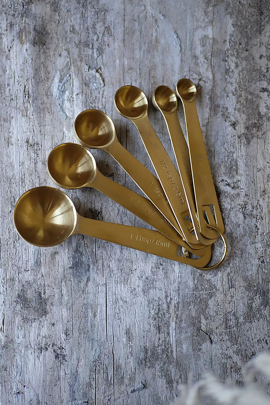 A set of seven brass coloured measuring spoons arranged in a fan shape on a rustic, weathered gray wooden surface.
