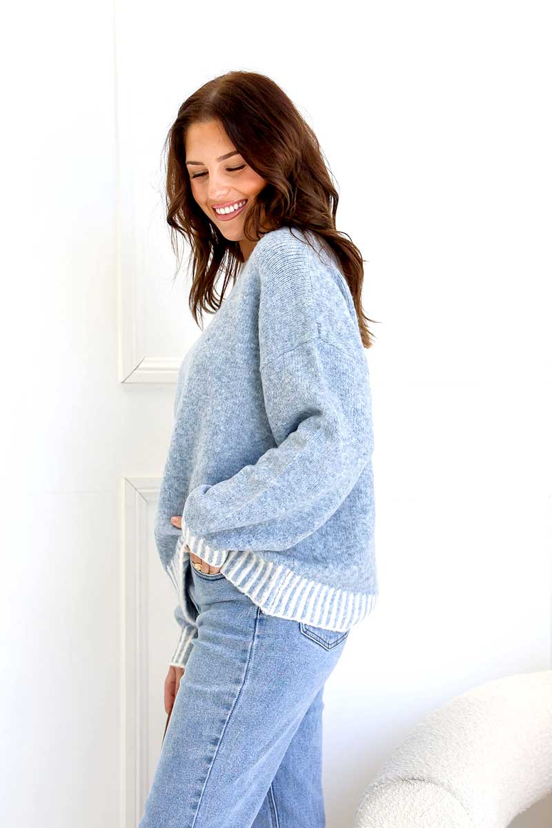 Woman wearing a Tilly blanket stitch Knit in blue and jeans, standing in a room with white walls, side on view.