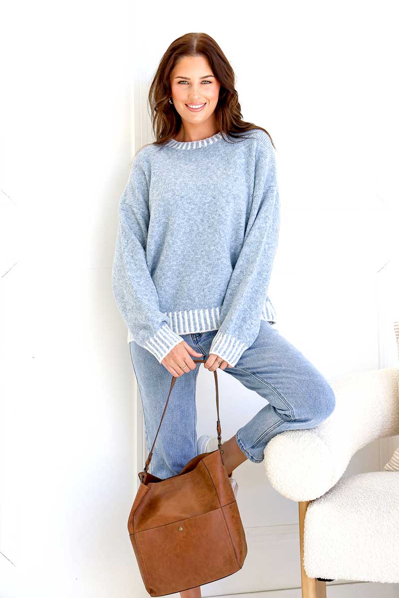 Woman wearing a Tilly blanket stitch Knit in blue and jeans, holding a brown leather bag indoors.