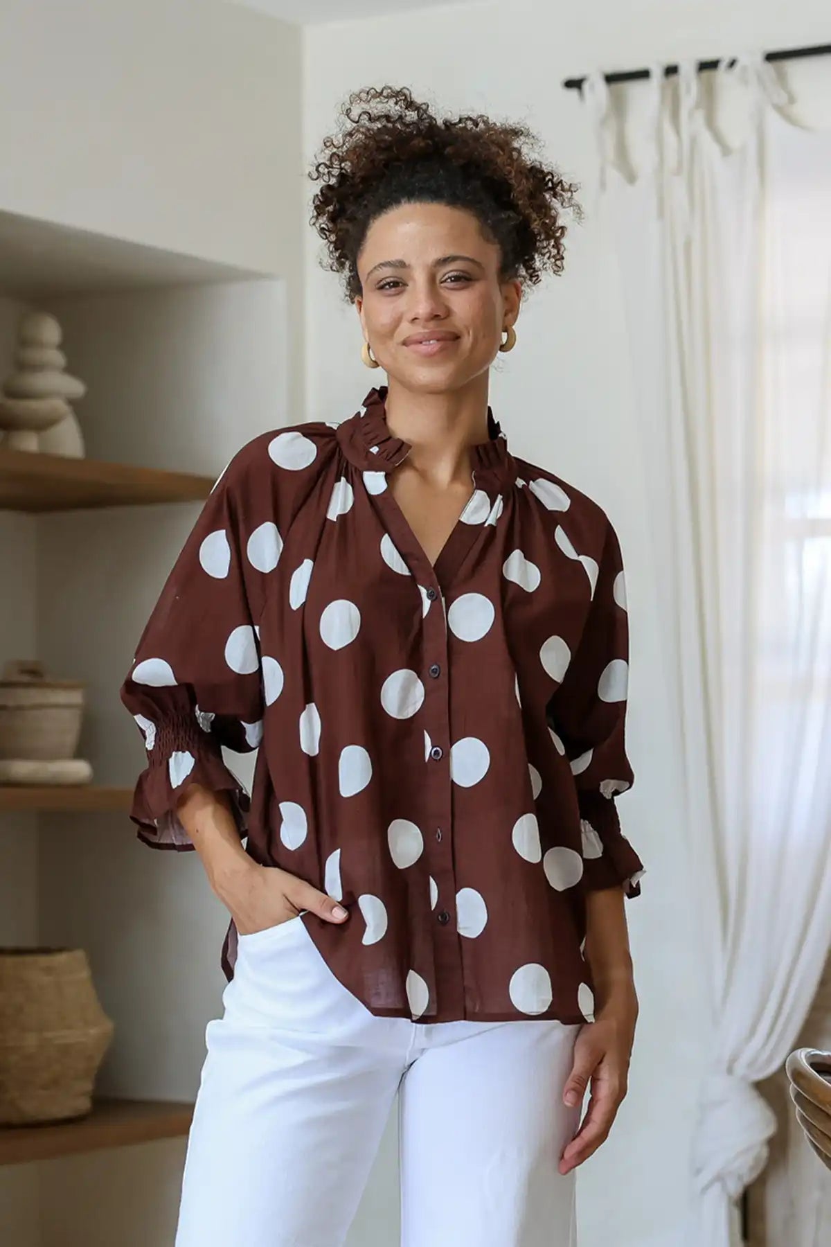Woman wearing a brown blouse with white polka dots and white pants indoors.