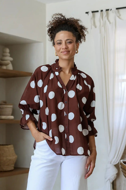 Woman wearing a brown blouse with white polka dots and white pants indoors.
