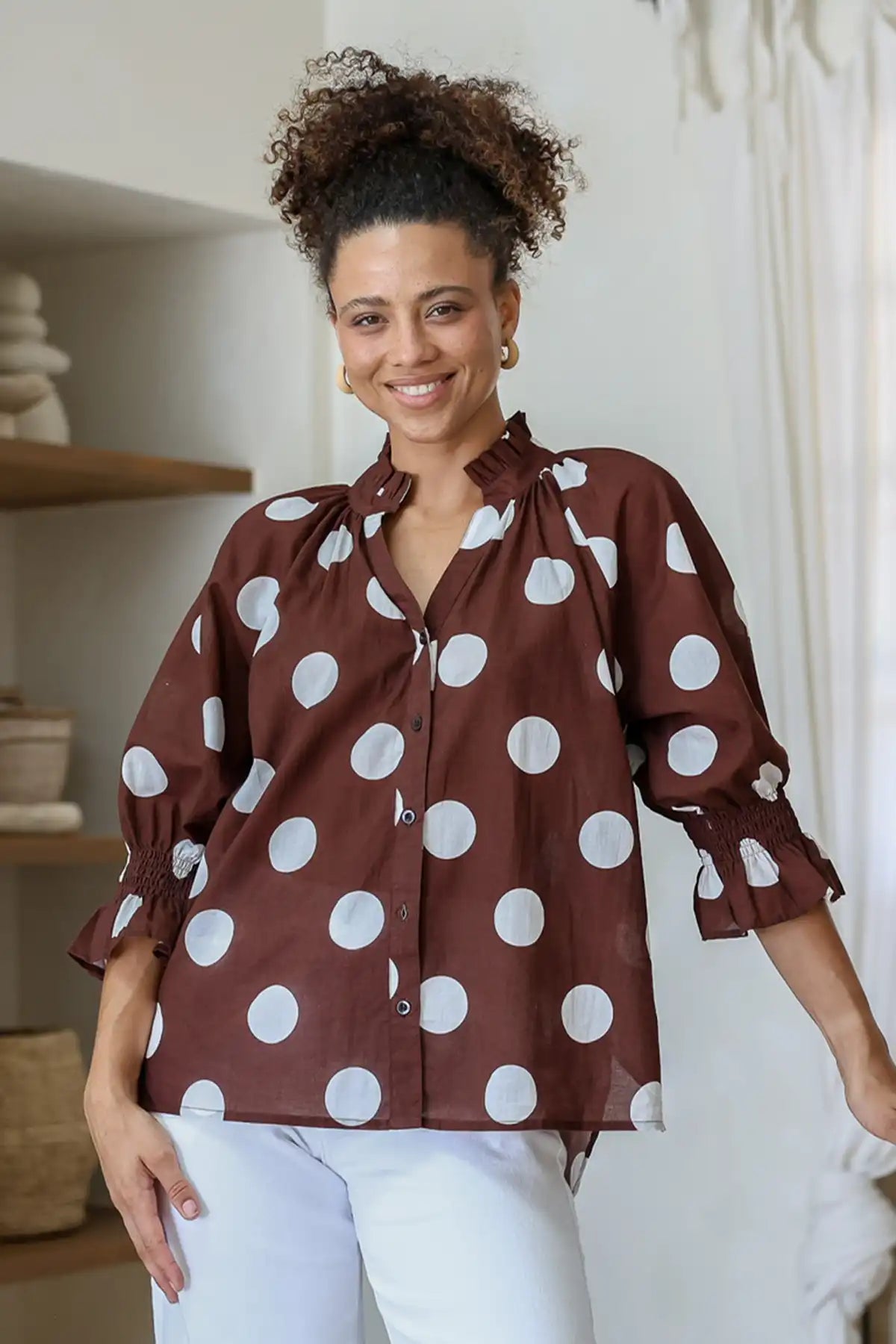 Woman wearing a brown blouse with white polka dots and white pants indoors.