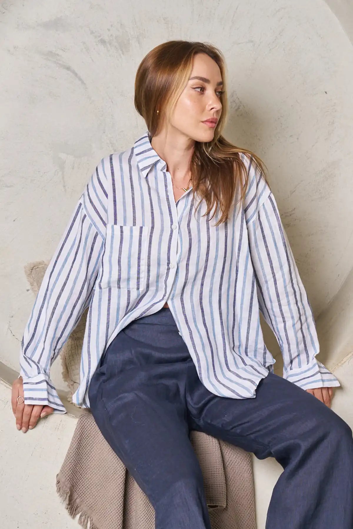 Woman wearing a striped shirt and navy pants sitting against a neutral wall.