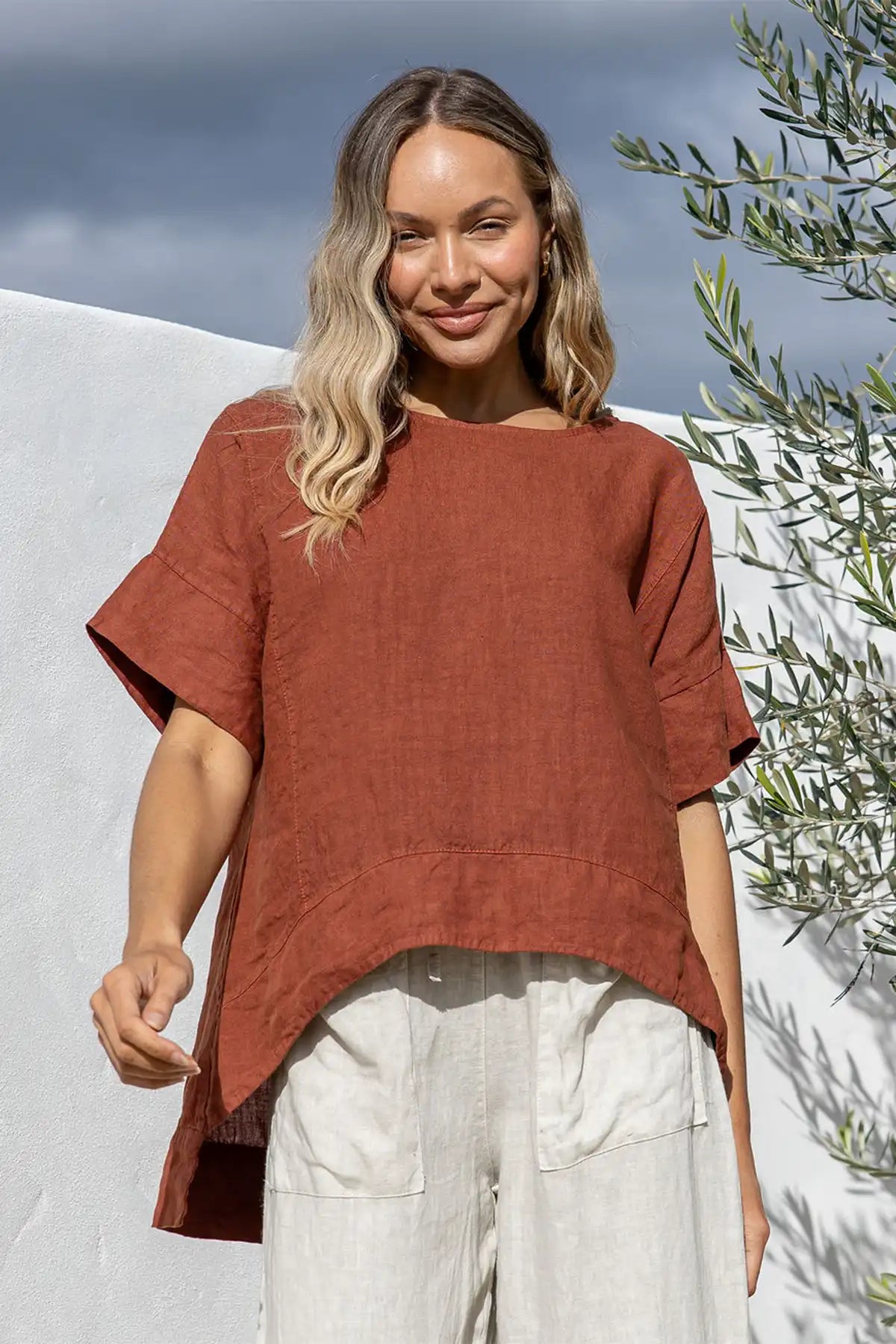 Woman wearing a Valentina Italian Linen Coco Top and light pants standing outdoors with a white wall and greenery in the background.