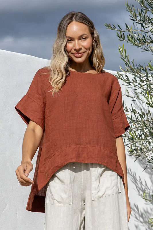 Woman wearing a Valentina Italian Linen Coco Top and light pants standing outdoors with a white wall and greenery in the background.