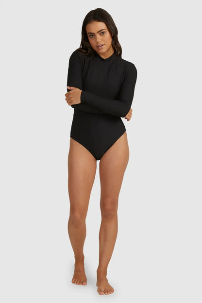 Woman wearing a Billabong Tanlines Bodysuit Rash Vest in Black long-sleeve  on a white background, full model view front on.
