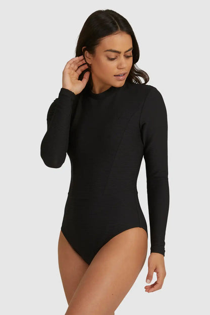 Woman wearing a Billabong Tanlines Bodysuit Rash Vest in Black long-sleeve on a white background, side view.