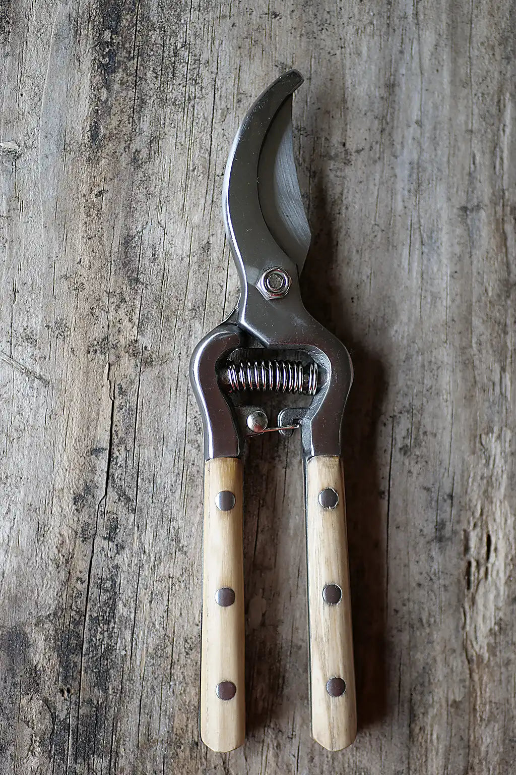 A pair of pruning shears with curved silver metal blades and light coloured wooden handles, shown closed on a rustic weathered wood surface.
