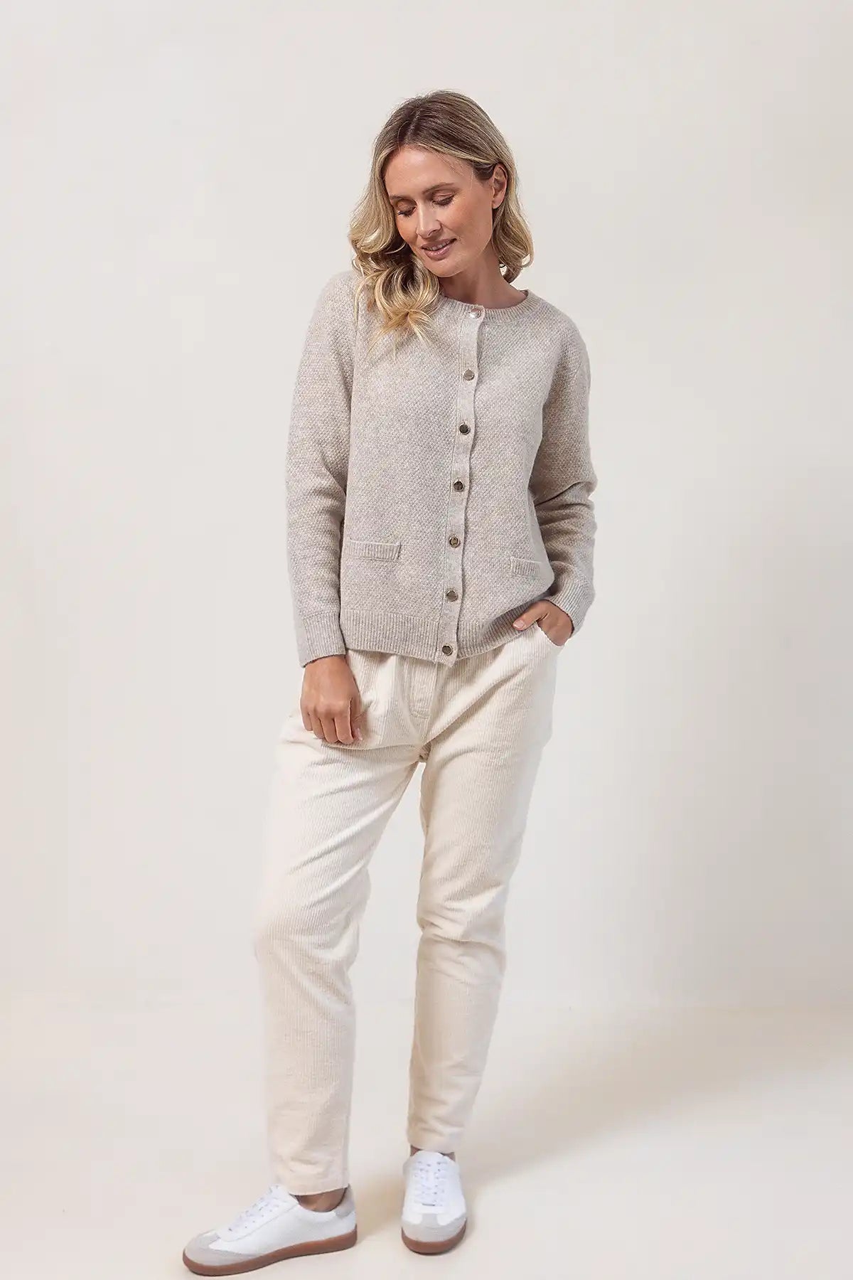 Woman wearing a wheat cardigan, white pants, and white sneakers on a plain background