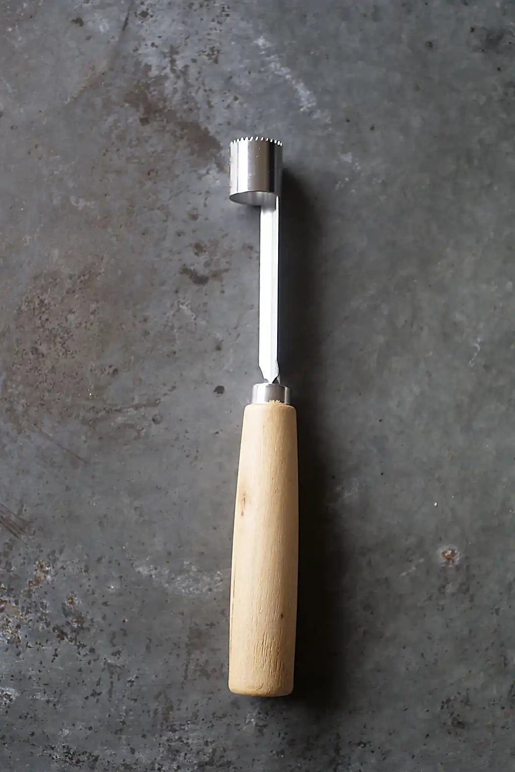 A meat tenderizer with a light-colored wooden handle and a stainless steel tip, placed vertically on a textured gray concrete surface.