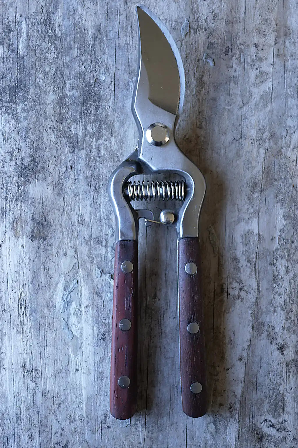 A pair of pruning shears with silver metal blades and dark reddish-brown wooden handles, laid flat on a weathered gray wood surface.
