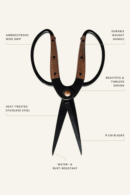 A pair of large scissors with black metal blades and walnut wood handles, shown on a light beige background with callout text labels describing features like wide grip, durable walnut handle, heat-treated stainless steel, 9 cm blades, and water and rust-resistant construction.