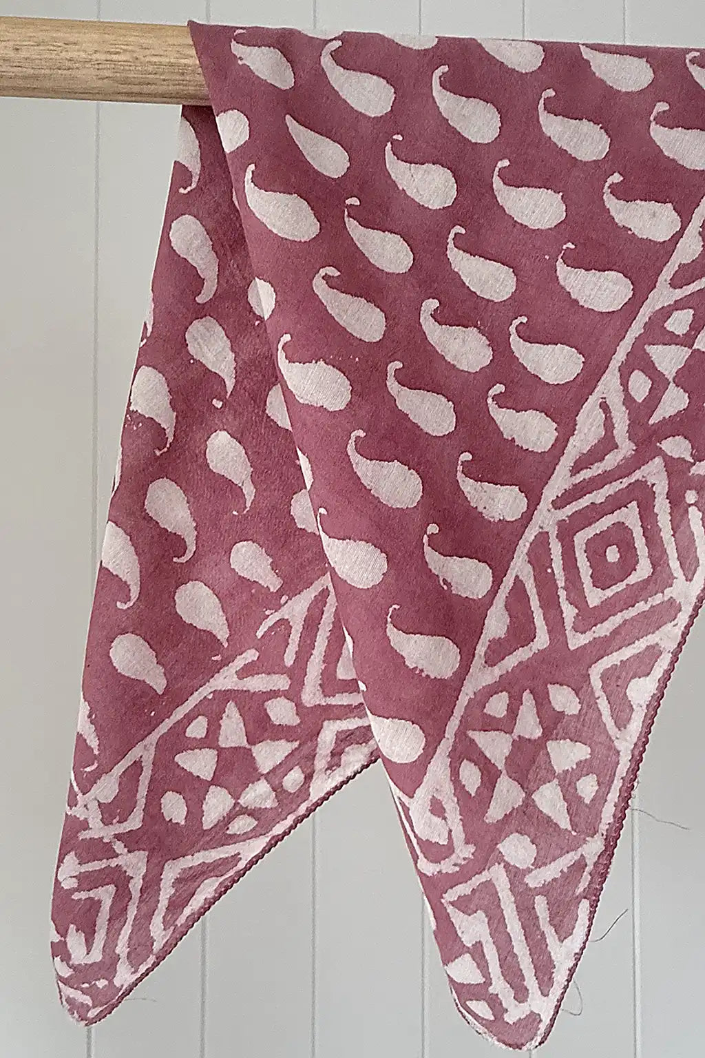 Pink cotton scarf with white patterns draped over a wooden rod against a white tiled wall.