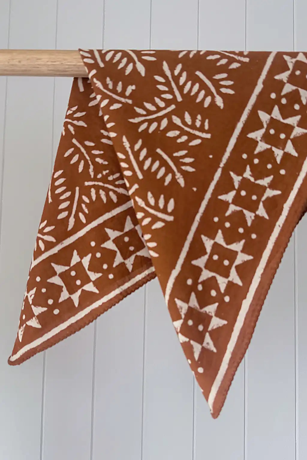 Cinnamon cotton fabric scarf with white star and leaf pattern draped over a wooden rod against a white wooden panel background.