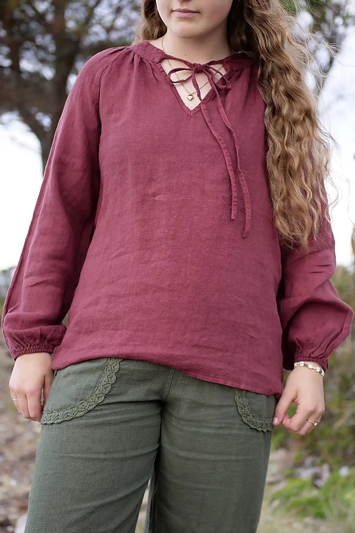 Person wearing a berry long-sleeve top with tassel tie-up front and green pants outdoors.