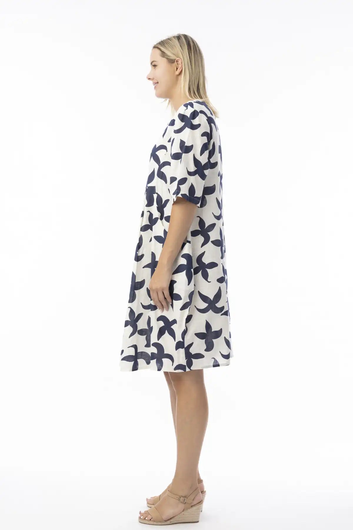 Woman wearing a white dress with blue floral pattern on a white background standing side on