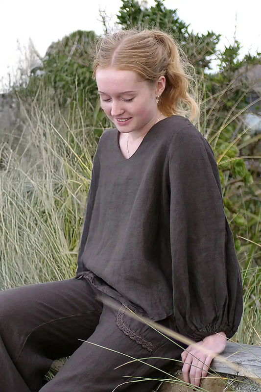 Woman in dark brown outfit sitting outdoors with greenery in the background