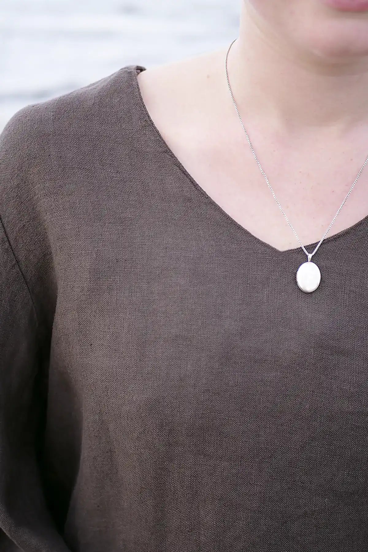 Person wearing a choclate linen top with silver necklace with a round pendant on a plain background