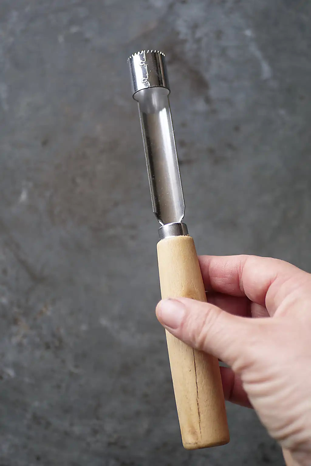 A hand holding a metal and wood kitchen tool against a textured gray background.