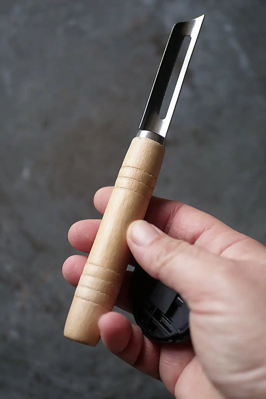 A hand holding a small utility knife with a light wood handle and a silver metal blade, with a black plastic cap or cover resting near the base of the handle against a dark textured background.